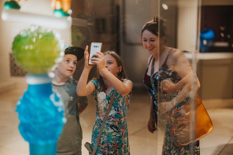 A mother and her two children look at a colorful indoor art exhibit. One of the children is using her phone to take a photo