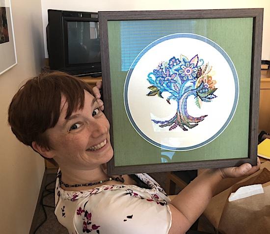 Jenn Poret is a white woman in her early 40s with short brown hair. She is posing sideways into the camera presenting a frame next to her face. In the frame is a cross-stitched tree in a mandala design within a white circle in front of a green background. Poret wears a solar system necklace and a white shirt with purple flowers. In the background, you can see a TV and a picture hanging on the wall.