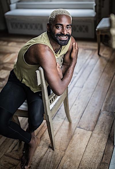 Jerron Herman, a black man in his thirties with short blonde hair, dark brown eyes and full beard, smiles at the camera from inside an ornate room, wearing green tank and black tights while leaning on the back of a chair. 