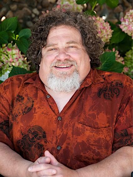 James LeBrecht is a white man in his sixties with curly brown hair and a gray and white goatee  the sides of his face are clean shaven. He is smiling, looking directly at us and is holding his hands together. James is wearing a short-sleeved, red, button-down shirt with black accents. Large flowers and green leaves are displayed within the background.