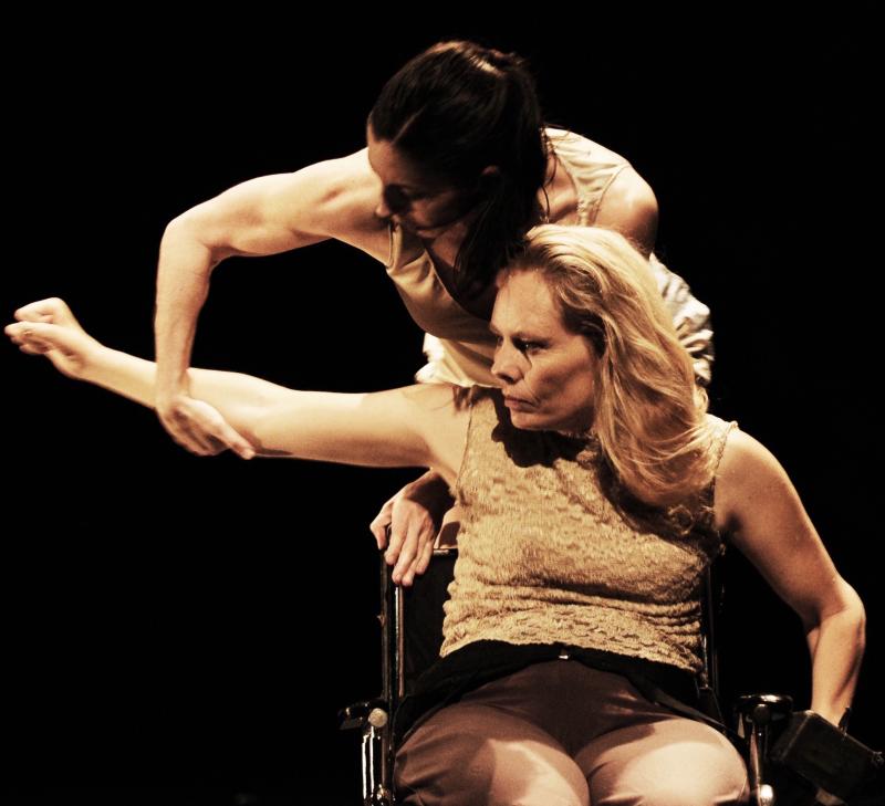 A sepia toned photo of Judith Smith in a wheelchair with her arm being held up by another person standing behind her. Judith is in her sixties, and is wearing a tan lace shirt and brown pants. Judith has long wavy blond hair. The dancer behind her is light in complexion and has dark brown hair pulled back into a ponytail. They are in front of a black background.