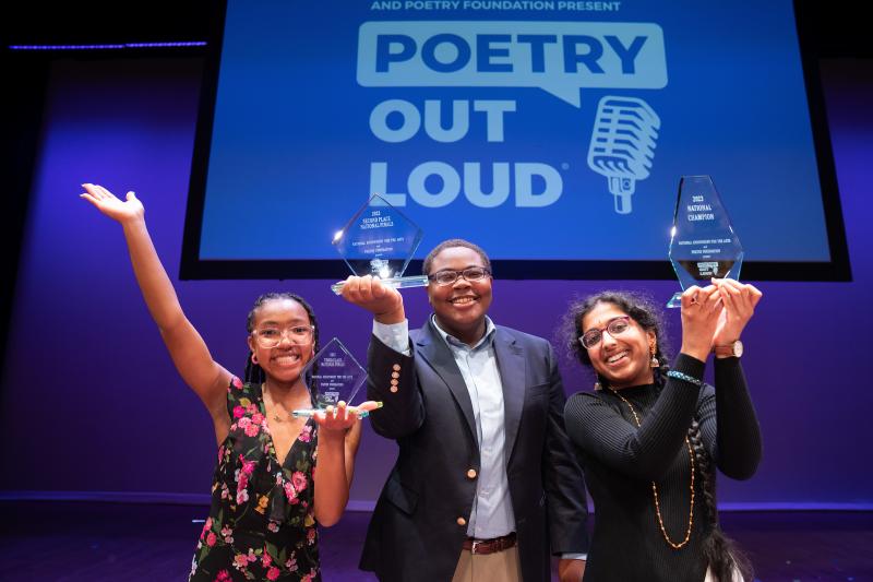 Photo of three students holding trophies in the air with the Poetry Out Loud logo on a screen behind them