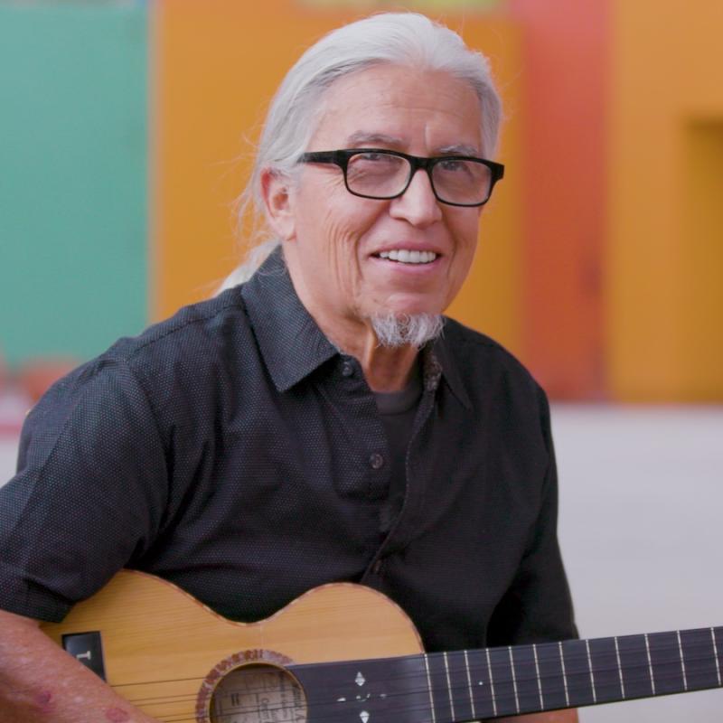 An older man holding a guitar smiles at the camera.