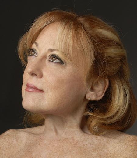 Mary Verdi-Fletcher is a white woman in her sixties with shoulder length reddish-blonde hair and freckles and brown eyes. Mary has decorated her eyes with black eyeliner that is paired with red lipstick. She gazes upwards, her neck and shoulders are bare, we see only her skin and is situated against a dark gray backdrop.
