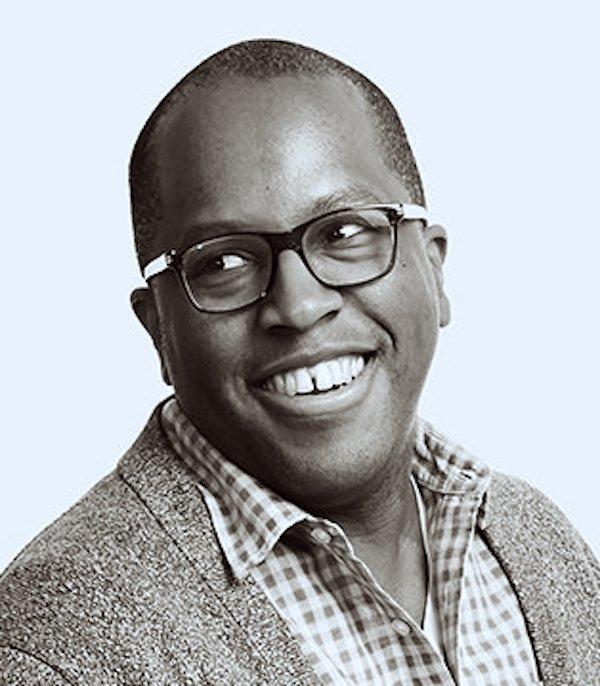 Headshot of a Black man smiling and wearing glasses, a checkered button-down shirt, with a jacket over the button-down shirt.