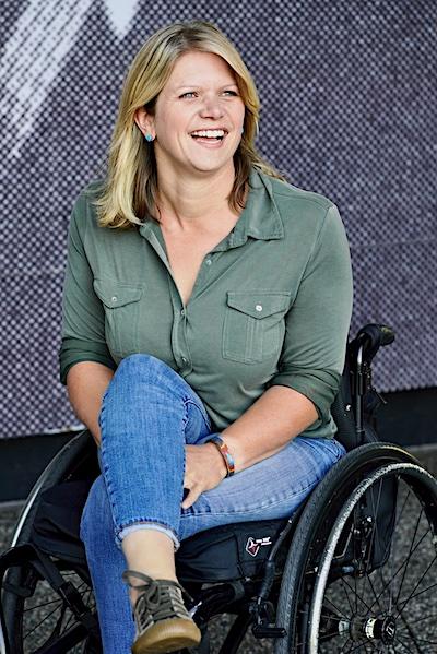 Regan Linton is a middle-aged Caucasian woman who uses a manual wheelchair. In this photo she has blonde, shoulder-length hair and is wearing a green collared shirt, blue jeans, and brown tennis shoes. She sits in a black manual wheelchair with silver hand rims, with a gray patterned wall in the background.  