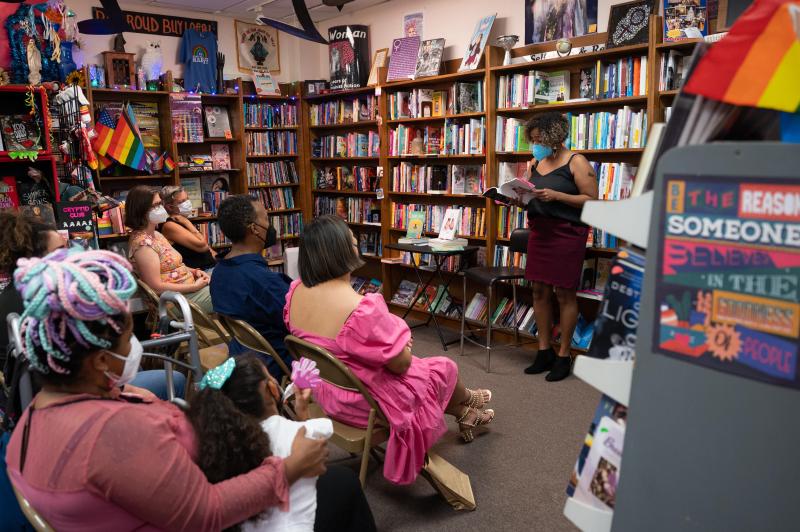 Woman in a mask stands in front of a wooden bookshelf filled with books and reads a book to an audience.