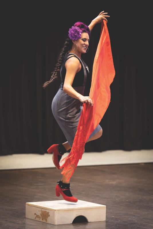 Vanessa Sanchez, who is a Mexican woman performs on a wooden platform. She is holding an orange scarf