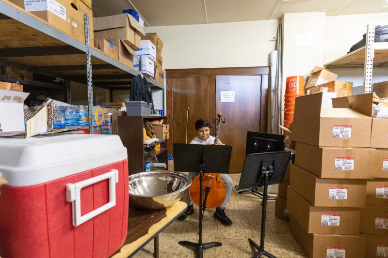 In a storage closet full of boxes and a large cooler, a boy sits behind several music stands, practicing the cello.