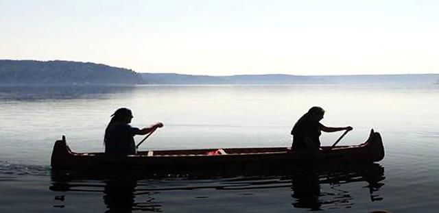 Two people paddling in a canoe on a large body of water with a hillside in the distance