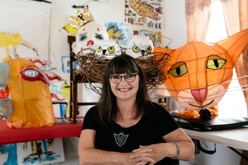 A woman sits and smiles at the camera surrounded by drawings and large paper lanterns that include a cat's head, a bird's head, a clown, and several owls in a nest.