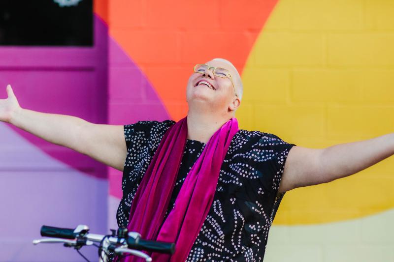 Petra Kuppers, a white queer disabled cis woman of size with yellow glasses, shaved head, pink lipstick and a black dotted top, smiles up to the sky, arms outstretched, embracing the world. Her mobility scooter’s handlebar is visible at the bottom of the image. She is in front of a multicolored wall: purple, pink, yellow and orange.
