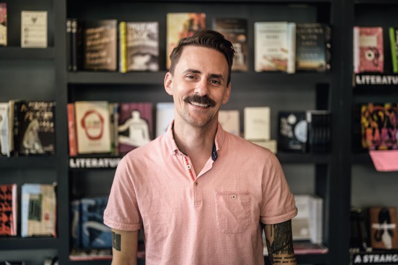 A white man with short brown hair and a moustache wearing a short sleeved light red collared shirt smiles in front of a bookshelf