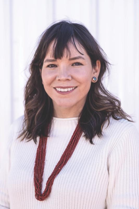 Heather Fleming, who is a Dine Navajo woman with long dark hair. She is smiling at the camera and wears red beads over a cream sweater