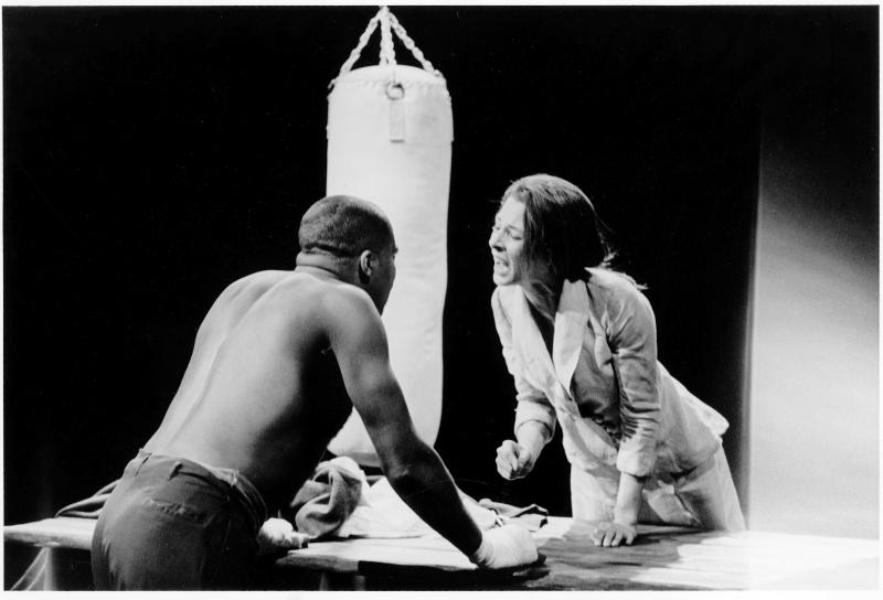 Shirtless man with short hair talking to woman in white suit over a table with punching bag in background. 