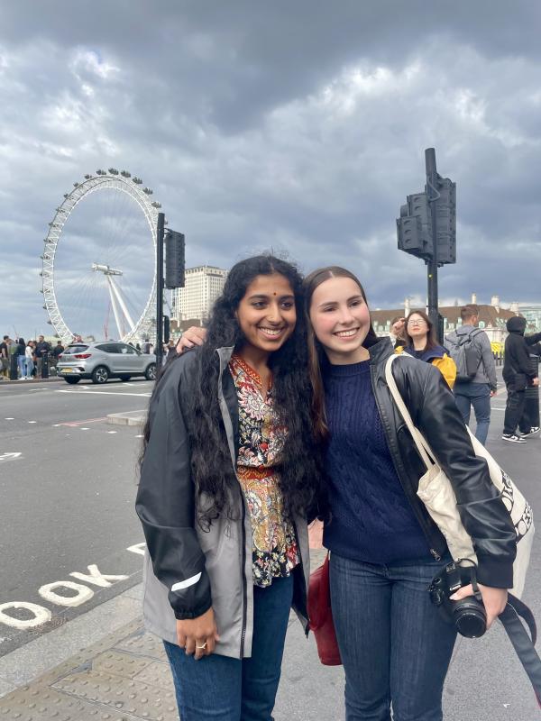 A young Indian woman and young white woman pose in front of the London Eye