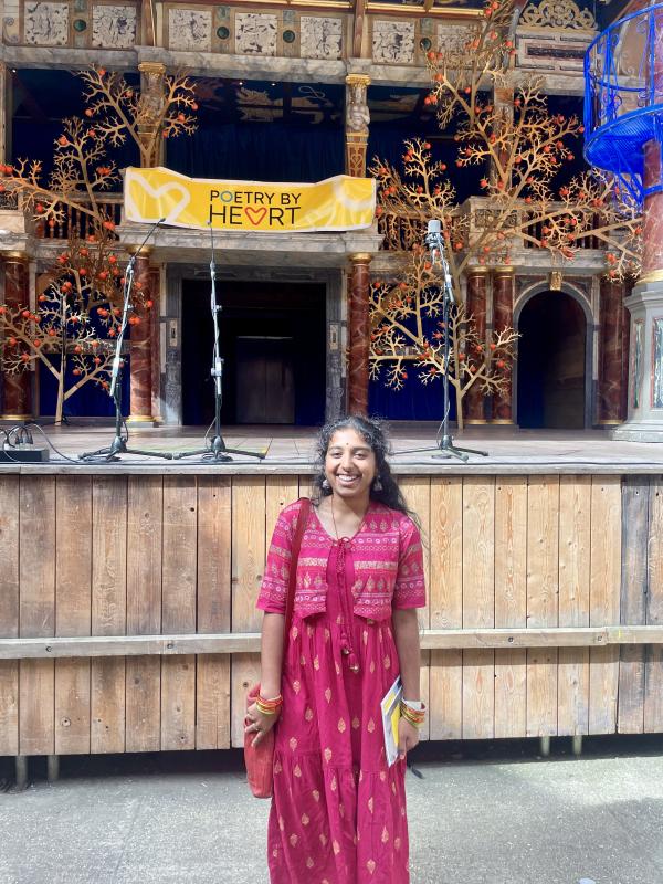 A young Indian woman stands in front of a stage with a banner that reads "Poetry by Heart" 