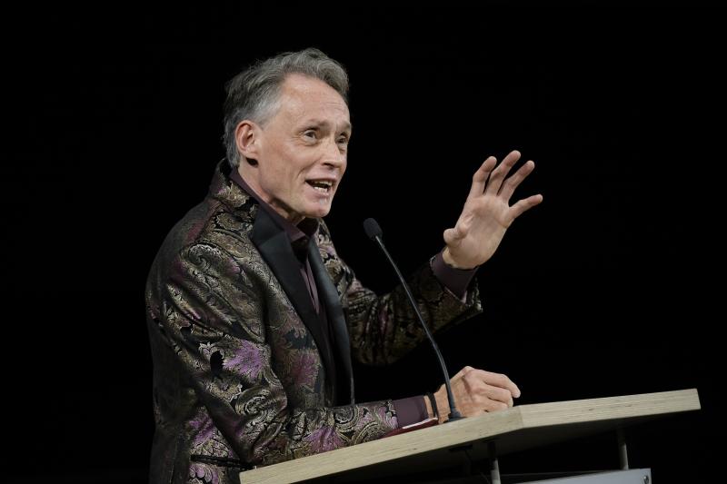 Lawrence Carter-Long, a white man with silverish blond hair, wearing a stylish purple, gold, and black tuxedo jacket and deep purple shirt, speaks into a microphone. One hand leans on the podium in front of him, the other is raised, mid-frame, punctuating the point he’s making.