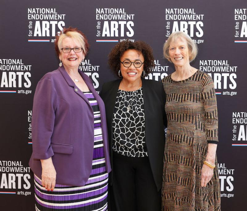 Three women pose in front of a banner with the NEA logo repeated.