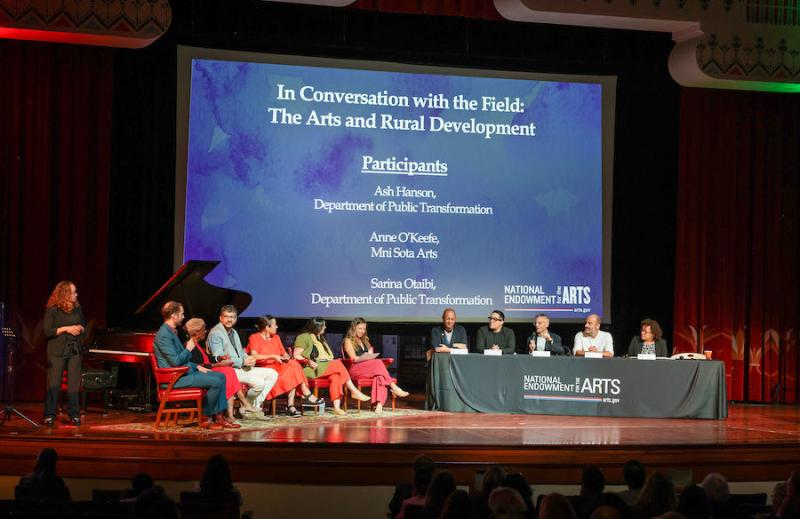 Six people are seated in chairs on one side of the stage. On the other side are five people seated behind a table with a tablecloth and the NEA logo on it. Behind them is a screen with the title of the panel and list of participants.
