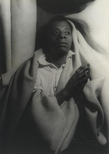 Black and white photo of a Black man with both hands clasped together with a white blanket behind him.