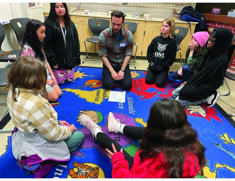 A man and several children are seated in a circle on a rug or mat showing a colorful map of the world and some of the animals native to the continents.