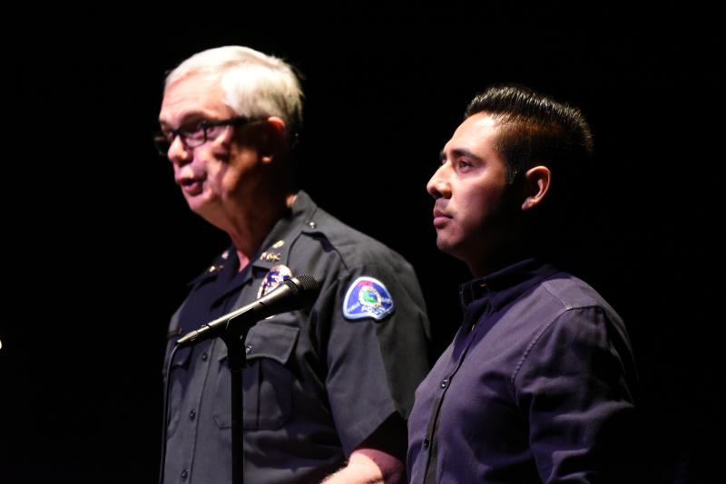 Older White man in a black police uniform (left) stands next to a Hispanic male (right) wearing a blue button-down shirt.