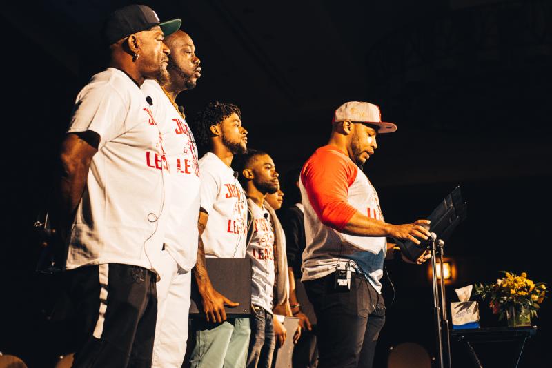Six black actors stand on stage (wearing red and white matching t-shirts) reciting monologues.