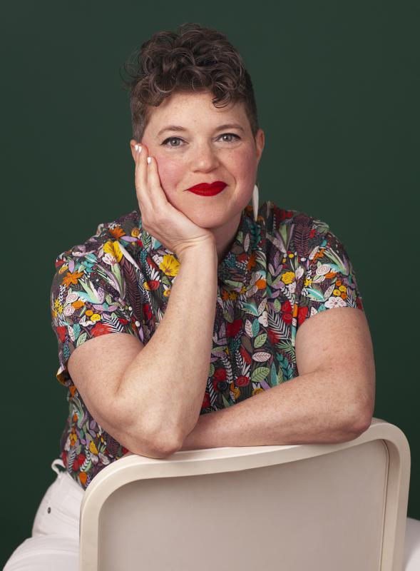 Headshot of a White woman wearing a multi-colored floral shirt with a solid green background behind her