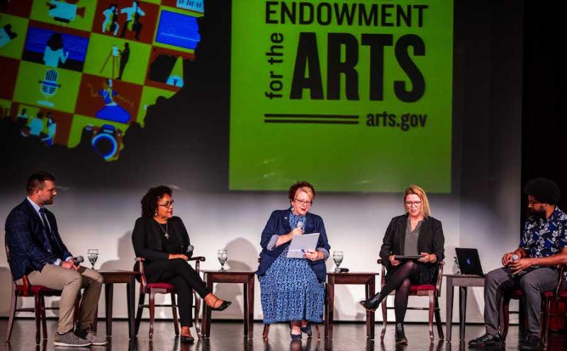 Five individuals sit in chairs on a stage, between them are small tables. Behind them is a large screen with the NEA logo and graphics representing a variety of art forms in the shape of the state of Ohio.