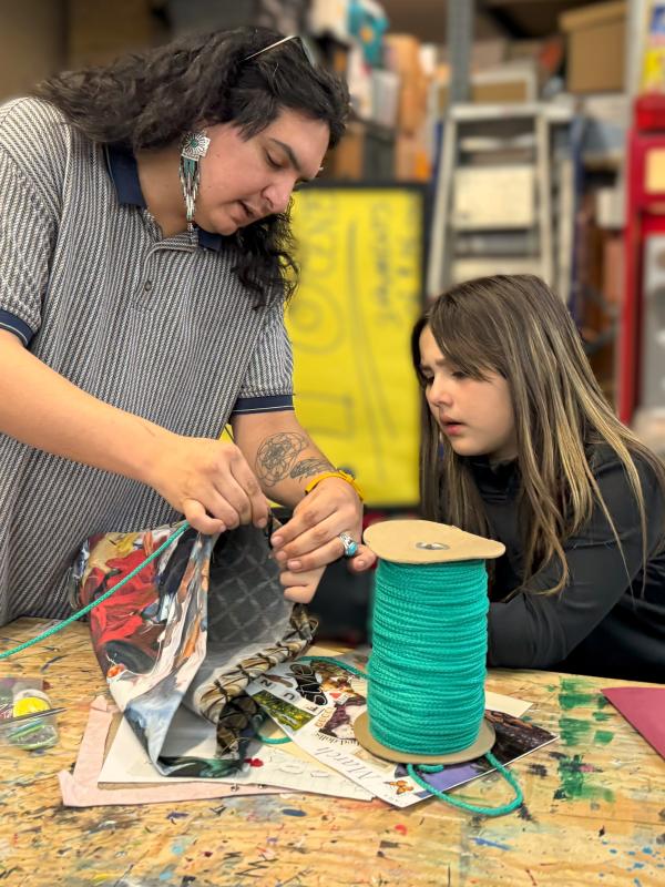 Male teaching artist (left) teaches a female student (right) are at a wooden table sewing.