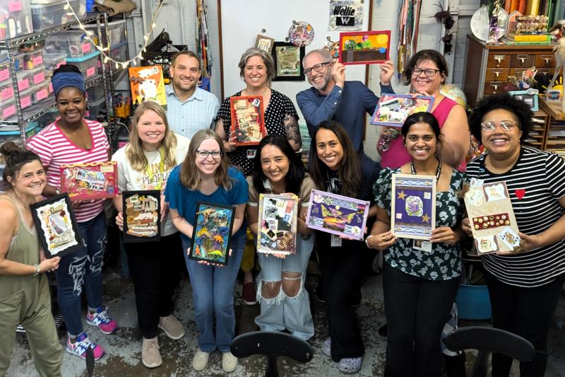 A group of teachers stand for a group photo, smiling and holding up their artwork.