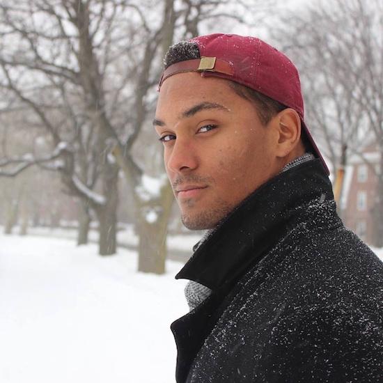 a young African-American man with a snow-dusted red baseball cap turned backwards and a black coat in a snowy background