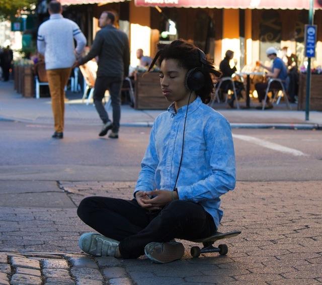 A ypoung in a meditation pose aittiing on a skate board while a number of people go about their business around him.