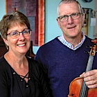 A man and a woman look towards the camera. The man hold a violin in his hand, a piano in the background.