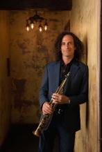 a full-shot of musician Kenny G wearing a dark blue suit and holding a soprano saxophone