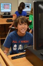 A young boy sits in the front of a computer with other students working on computers in the background.