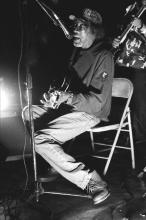 A black and white photo of an older man wearing a baseball-style hat, sitting on a folding chair playing the guitar and singing into a microphone. You can see another person in the background also playing music