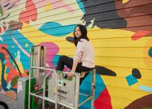 Asian women on scaffolding in front of mural she is painting. 