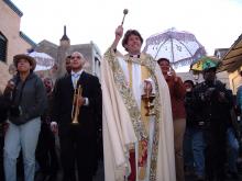 Man with trumpet next to priest blessing crowd in parade down street. 