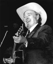 Man with gray beard in white hat playing a guitar. 