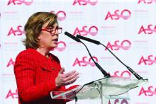 A woman speaks at a podium, a step-and-repeat banner is behind her which shows the logo for the Arkansas Symphony Orchestra.