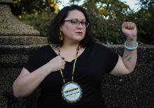 a Native woman with short wavy hair wearing glasses and a large beaded pendant that says Crushing Colonialism