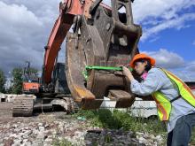 A person wearing an orange hard hat and a yellow safety vest attaches a camera to the claw of a large construction excavator. The claw has a green ribbon around it, and the person is leaning forward, peering through the camera's viewfinder.