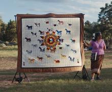 Susan Hudson standing outdoors with a large quilt displayed on a frame