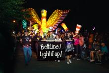 People lead a parade at night, holding a large sign that says "Beltline! Light it up!" They, as well as several others, are wearing tall hats that look like illuminated paper lanterns. Behind them is a large illuminated paper lantern of an orange and yellow bird.