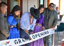 A small group of people stand behind a banner reading "Grand Opening". A man in the center is cutting the banner with large golden scissors.