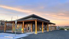 The exterior of a one story building with a colorful mural painted on one side. There are tables and chairs situated under an overhang supported by raw wood columns. The sky is filled with soft pastel pinks, blues, and yellows in what looks to be either sunrise or sunset.