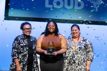 Three women stand smiling on a stage as confetti fall around them, the young woman in center holds a trophy 