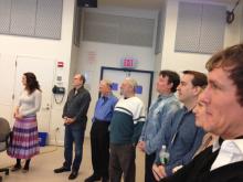 Group of people standing in a semi-circle in a beige room watching something to the left that is off-camera. 